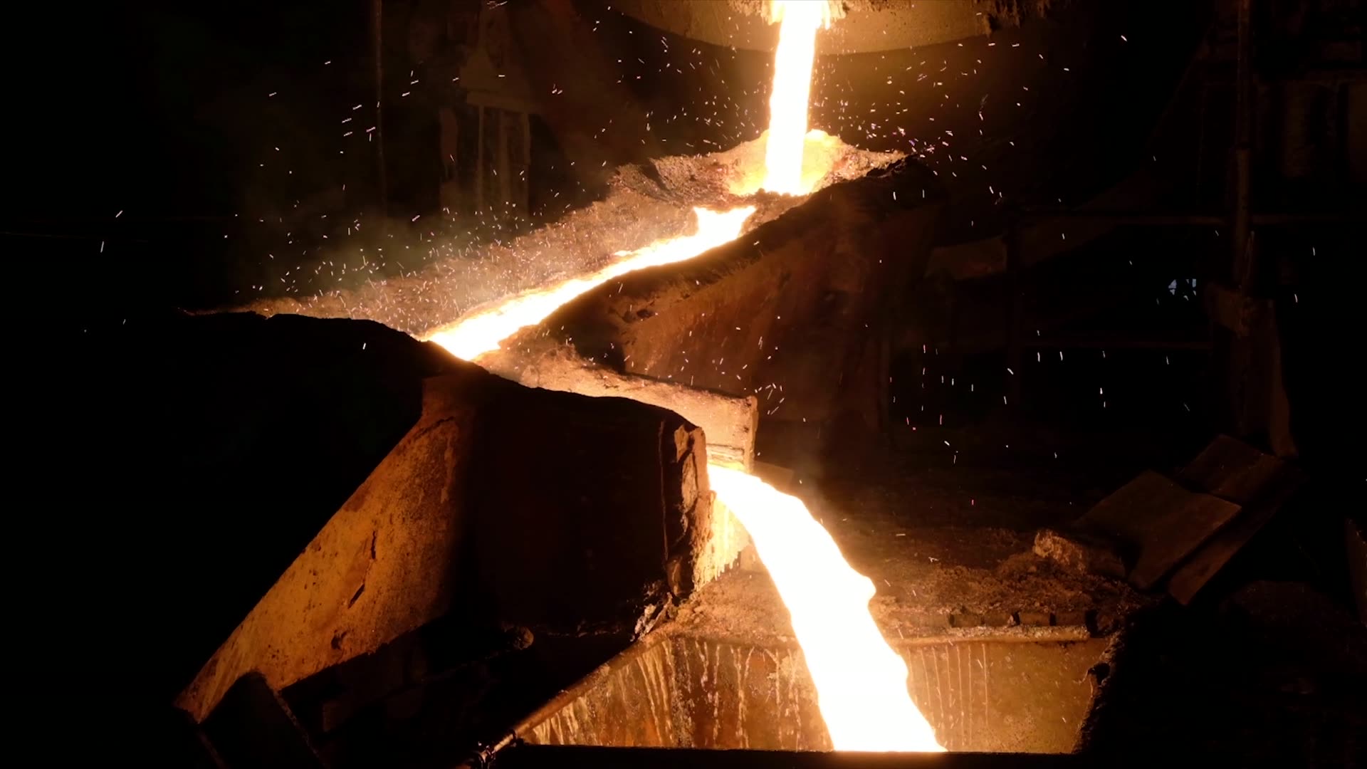 Molten steel pouring from a ladle in a mill, throwing orange light across the casting floor.